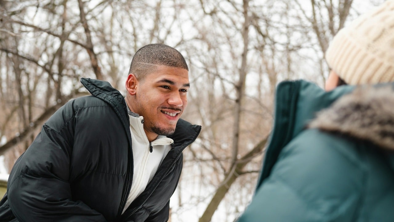 A smiling man in a black puffer jacket is looking and talking to a person in a green jacket and knit hat in a snowy outdoor setting.