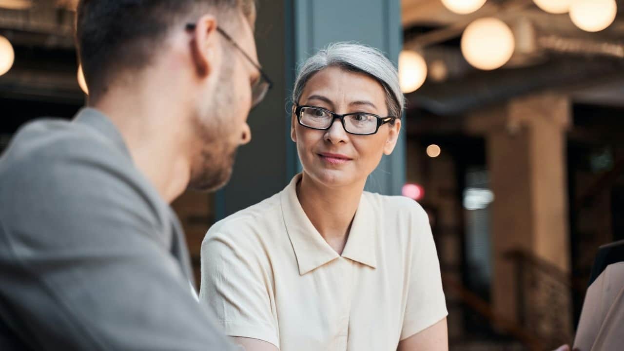 A woman with glasses and grey hair looks at a man in an indoor setting.