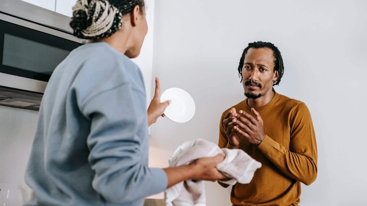 A woman is holding a plate and towel, gesturing while speaking to a man who looks distressed.