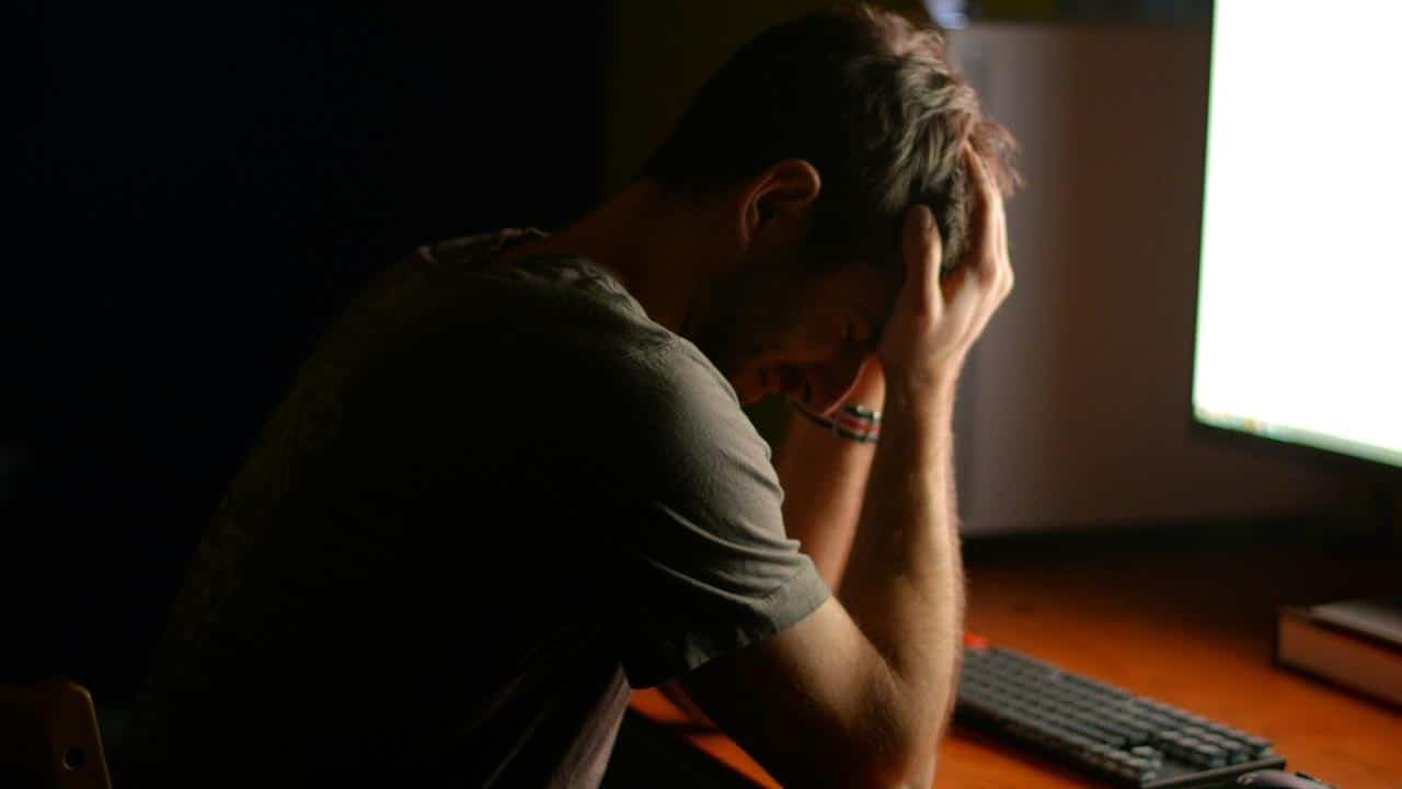 A man sitting at a desk holding his head in frustration near a computer.