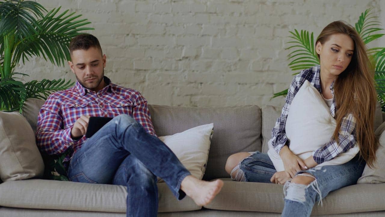 A man is using a tablet while a woman sits on the same couch looking sad, with both ignoring each other