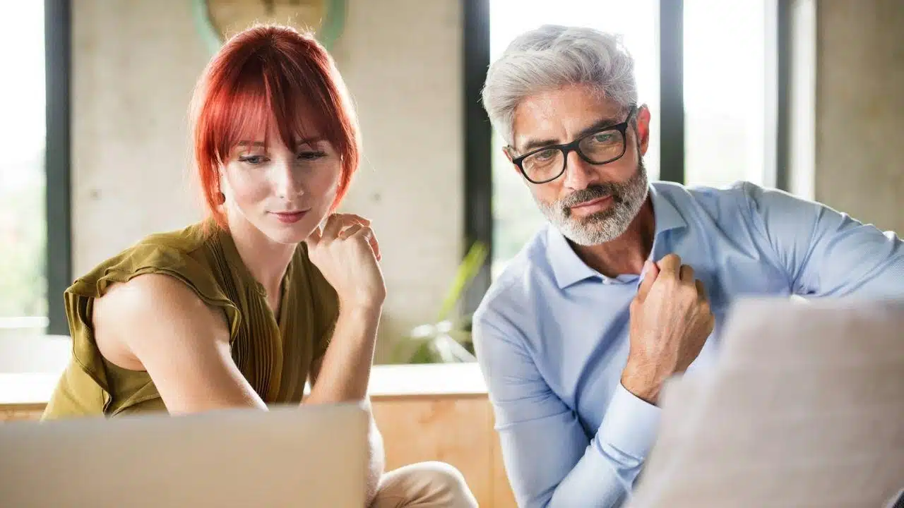 A younger woman and an older man with glasses look down at a laptop screen together.