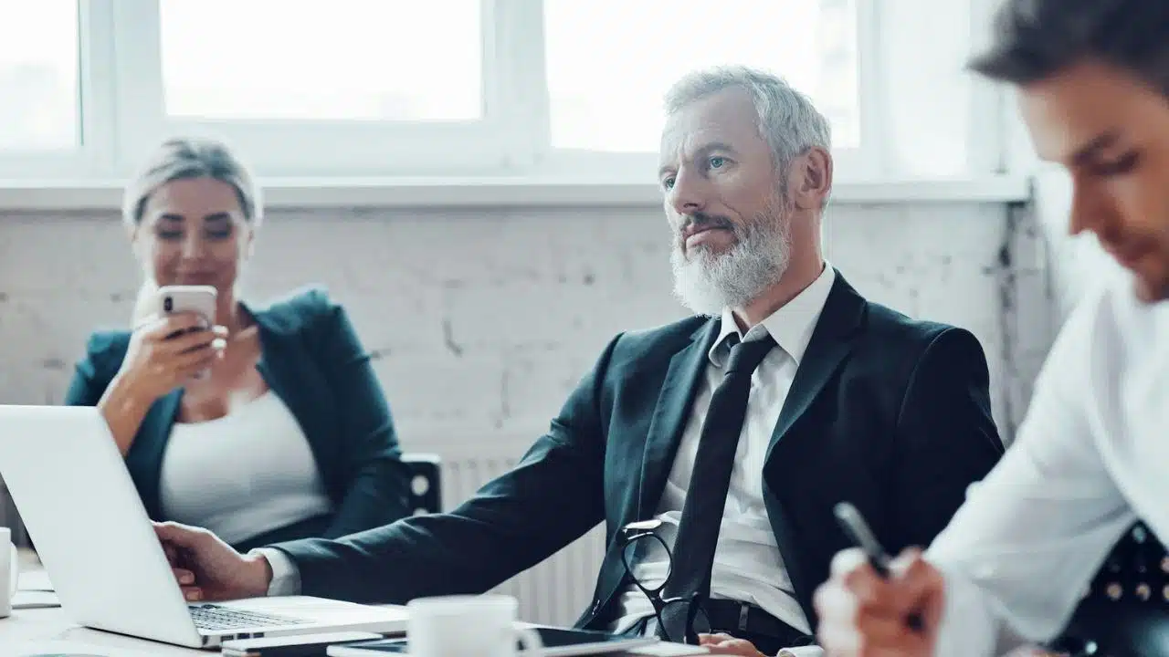 Bearded senior man in a suit looking to the side while sitting at a meeting table with a laptop.