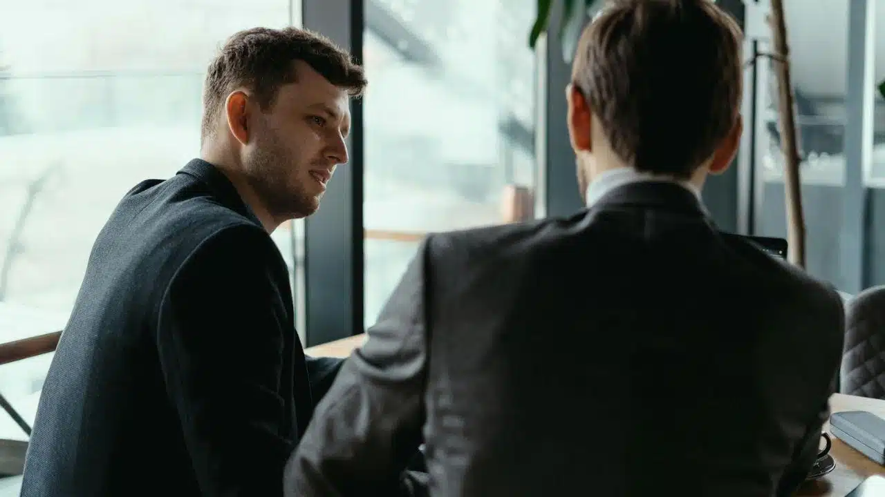 Man in a black jacket leaning in to talk to another man in a gray suit jacket.