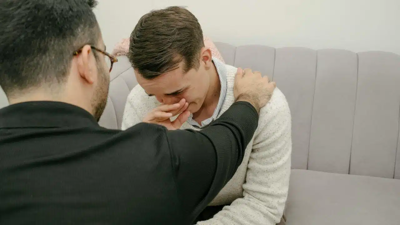 Man comforting another man who is crying and covering his face with his hand on a gray sofa.