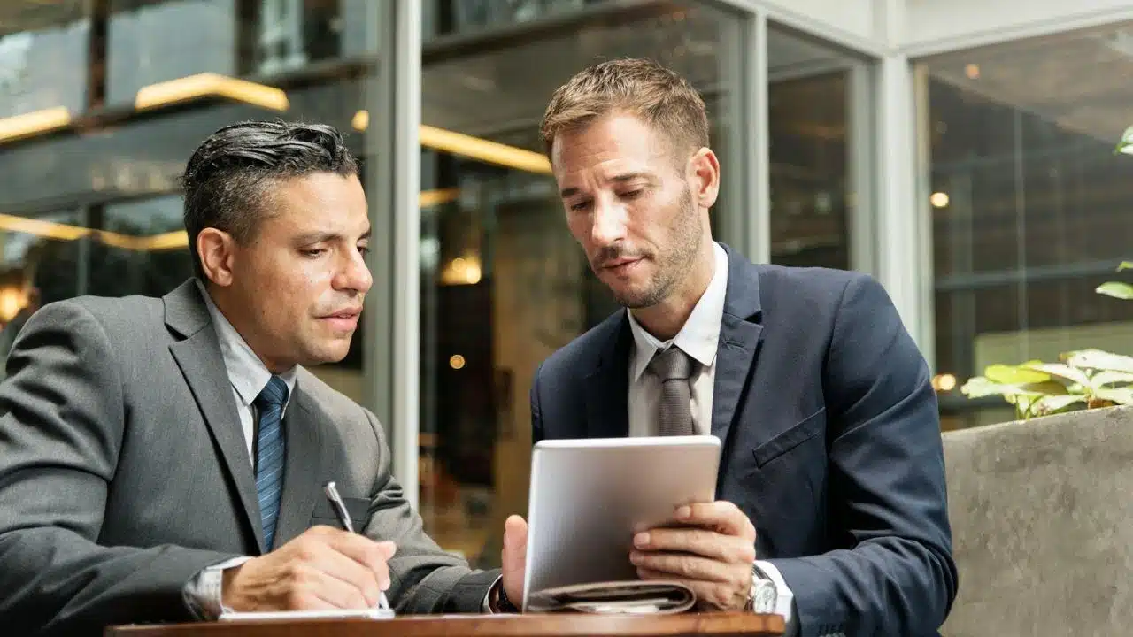 Two businessmen in suits look intently at a tablet while one writes on a notepad.