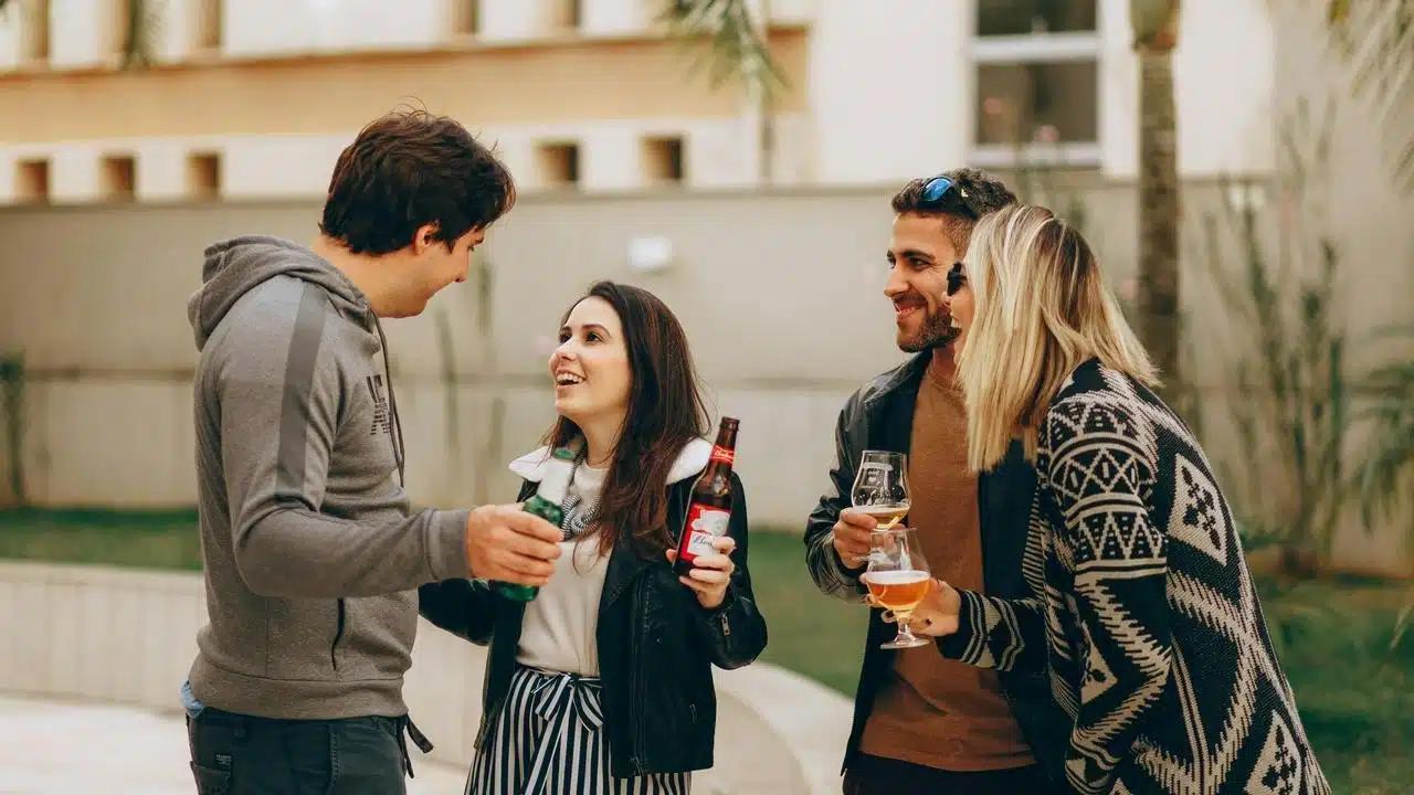 A group of friends stand outdoors talking and holding drinks.