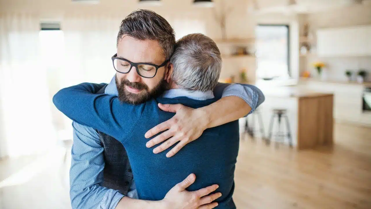 Smiling man with glasses and a beard embracing a gray-haired person in a bright, modern kitchen and living area.