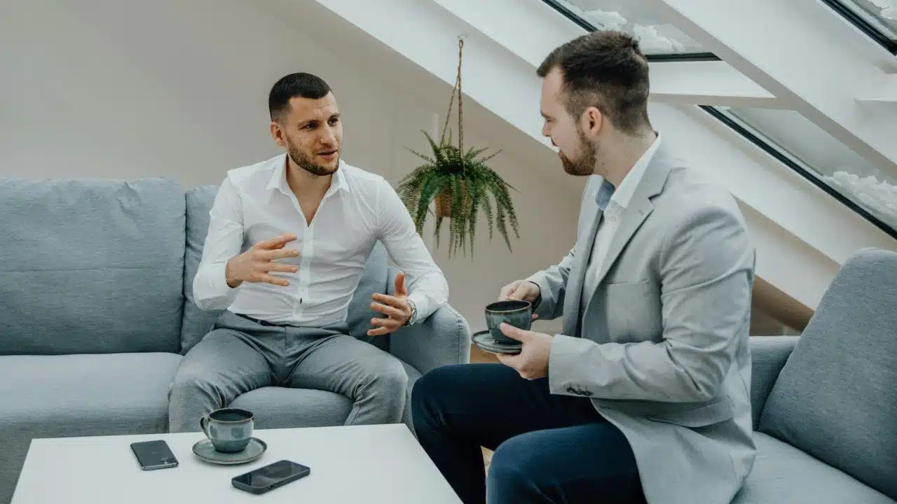 Two men are having a serious conversation while seated on a light blue couch indoors.