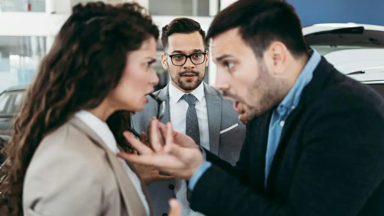 Man in a gray suit and glasses looks on with concern as a man and woman angrily face each other.