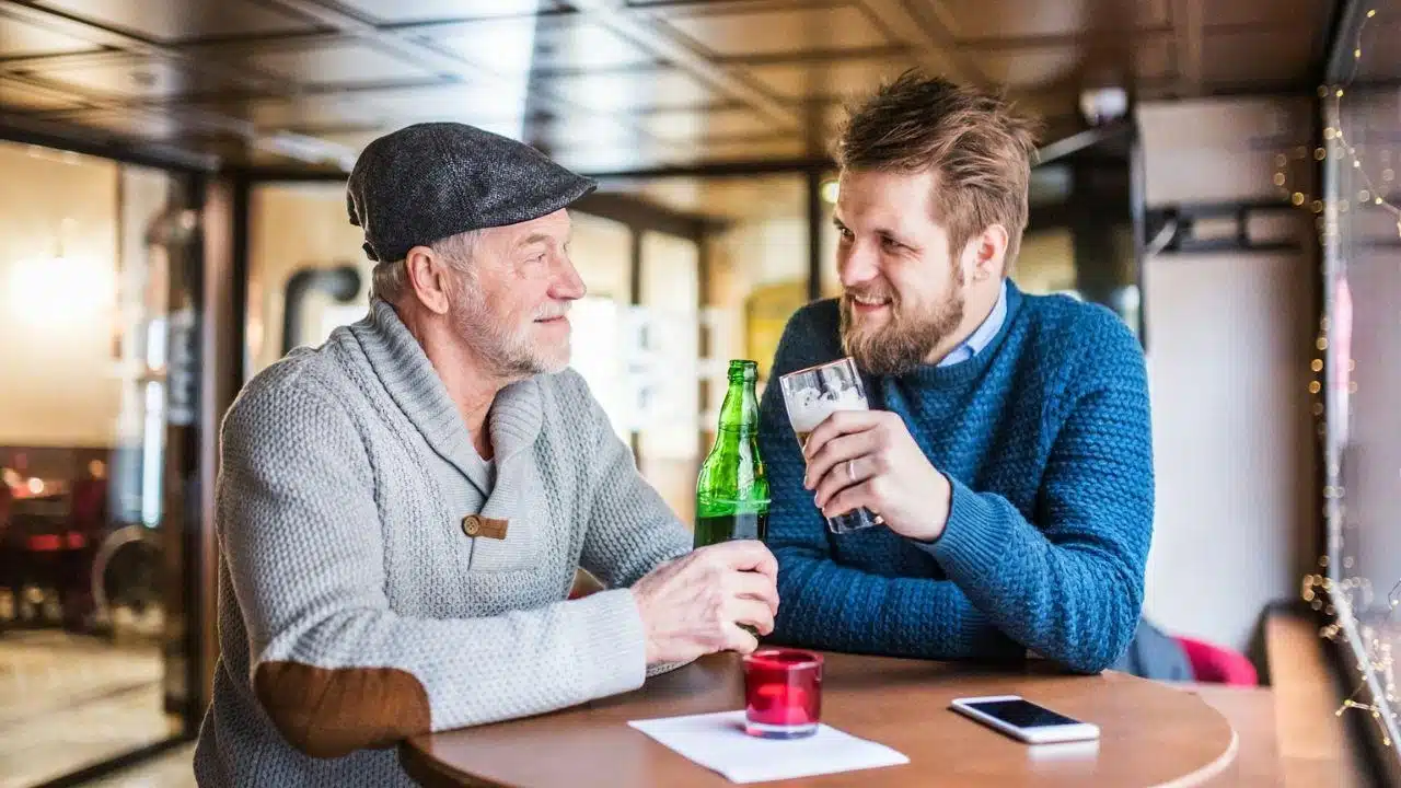 Two men, one older and one younger, talk while drinking in a dimly lit bar or pub.