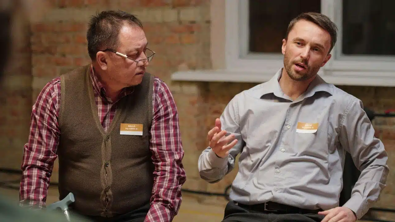 Two men with nametags in a casual setting, one talking and gesturing while the other listens.