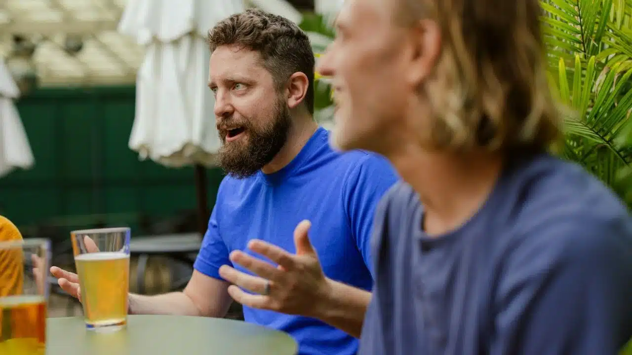 Two men talking at an outdoor table with drinks, one wearing a blue shirt.