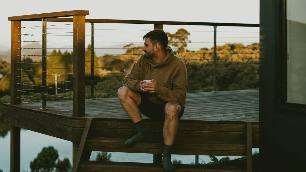 A man sits alone on a wooden deck, holding a cup and looking out at the scenery.