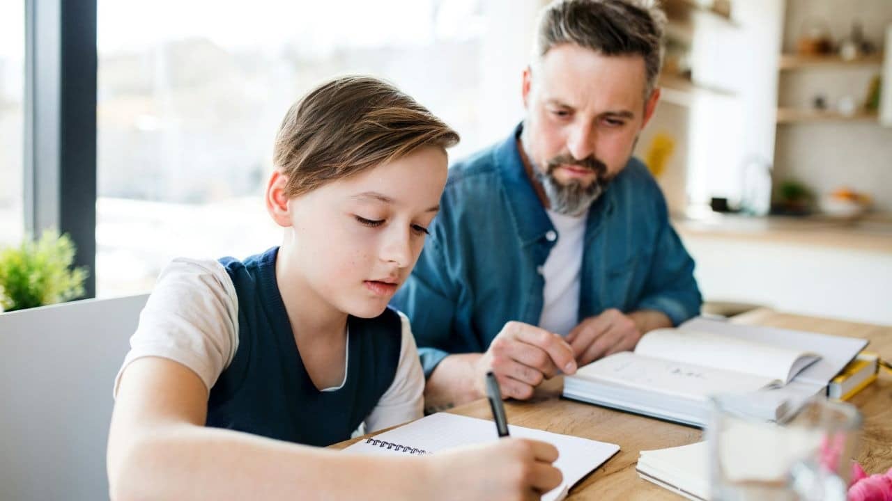 A father with a beard sits at a table helping his young son with his homework.