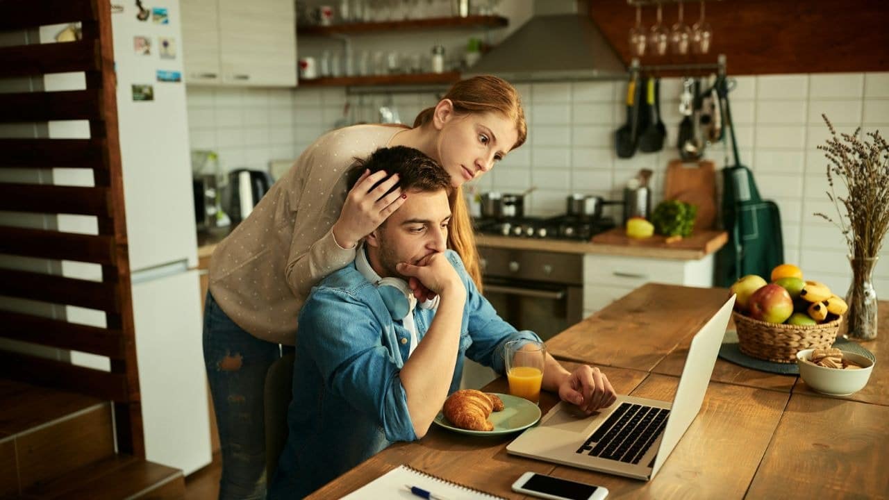 A worried woman comforts a stressed man as he looks at his laptop in the kitchen.