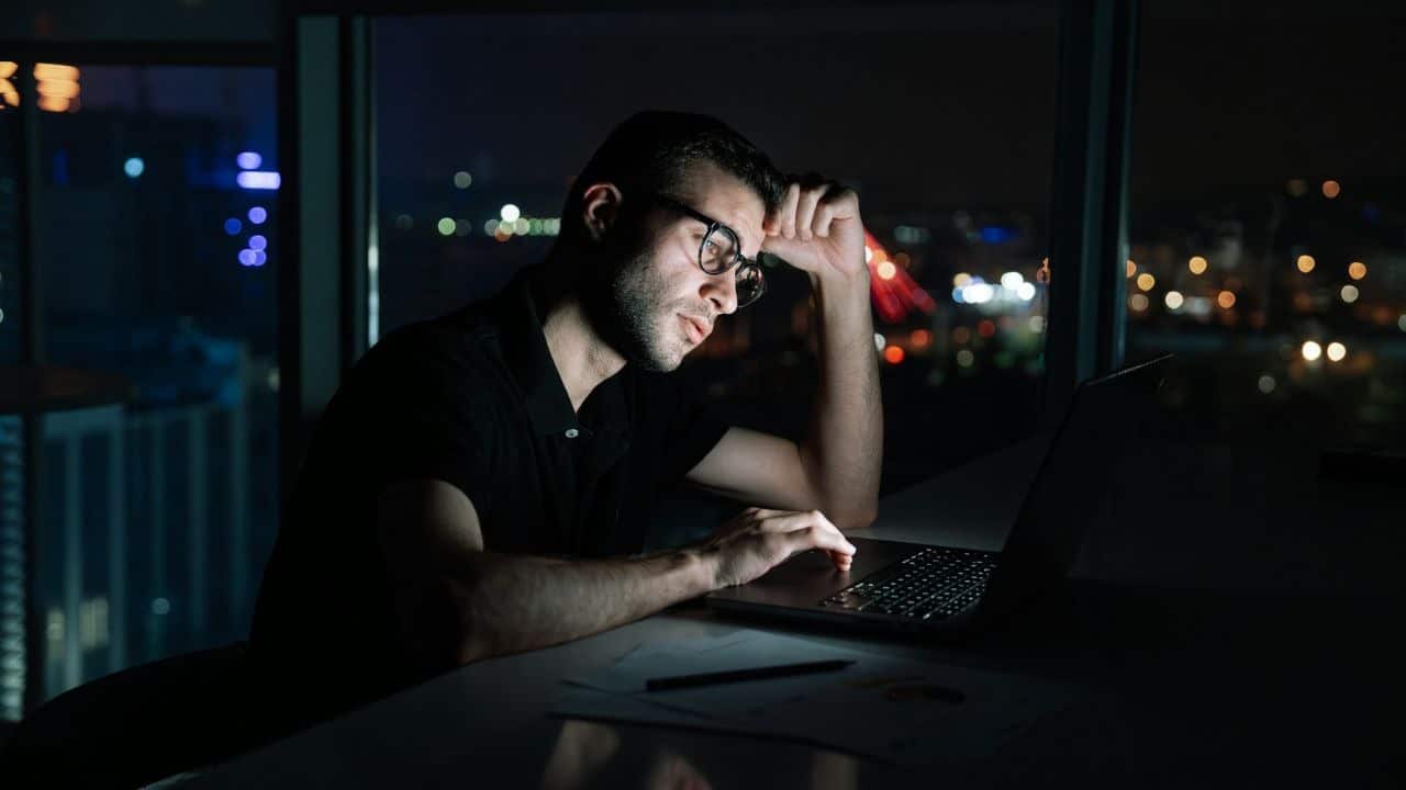 A tired man in glasses works late at night on his laptop in a high-rise office.