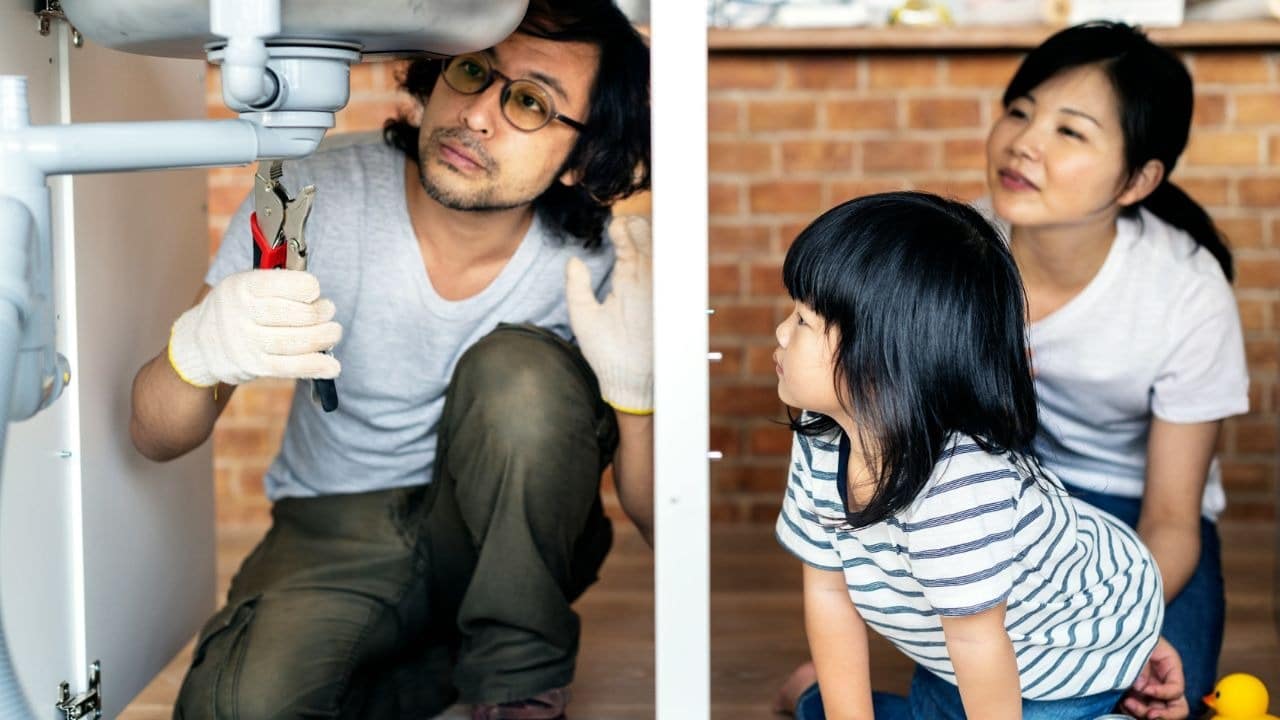A father wearing glasses and gloves works on a sink pipe with a wrench while his daughter and wife watch him.