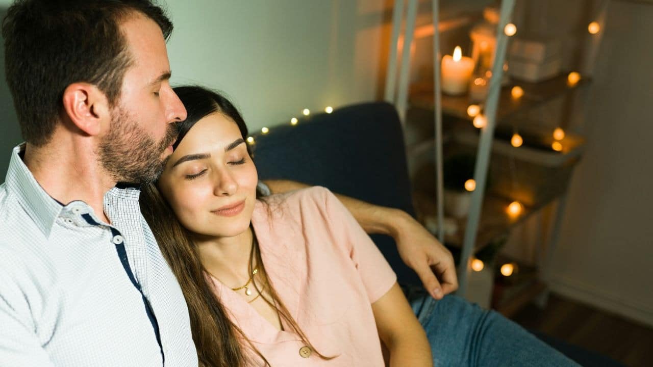 A couple relaxes together on a couch, lit by string lights and a candle.