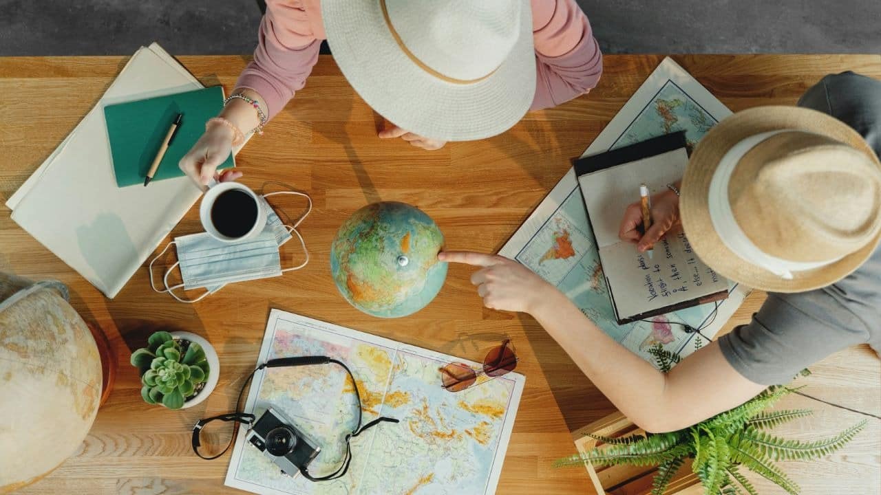 An overhead view shows two people with hats planning a trip with a globe and maps.