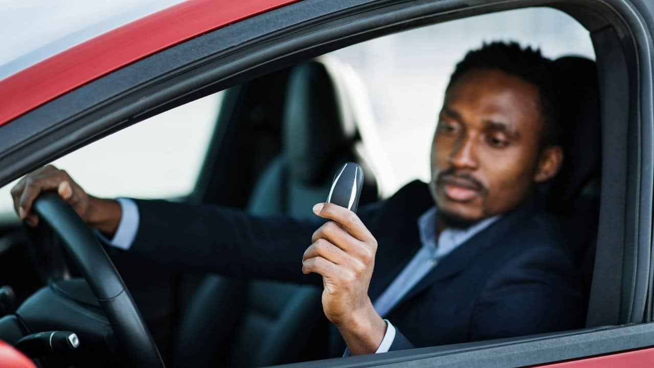 A man in a suit sits in a red car, holding up the car key and looking at it.