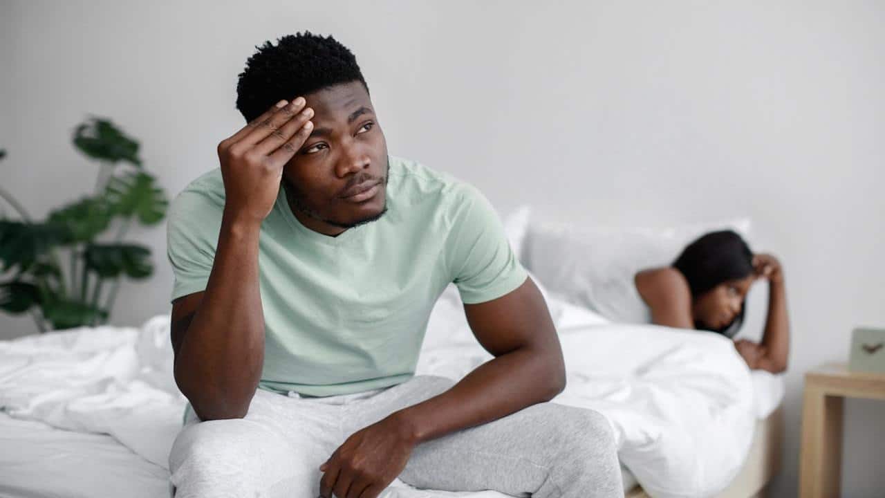 A man sits on a bed looking stressed with his hand on his forehead, while a woman lies facing away from him in the background