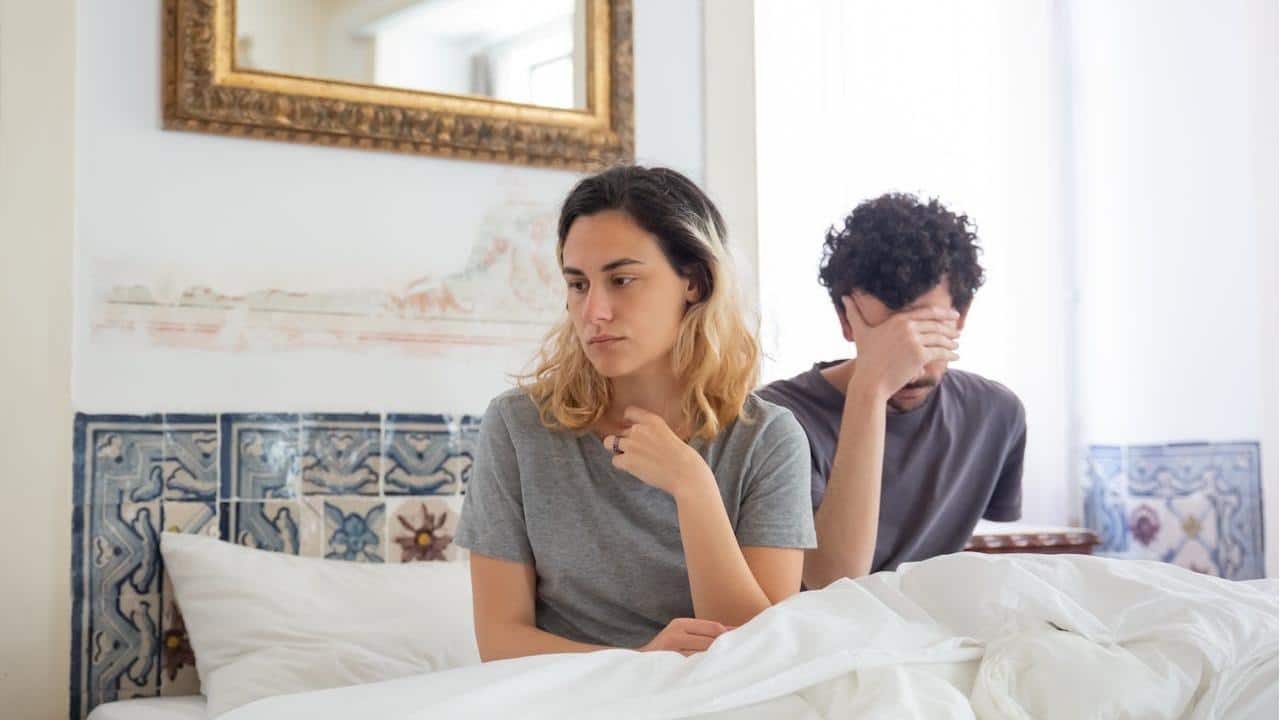 A couple sits on a bed after an argument; the woman looks sad while the man holds his hand over his face.