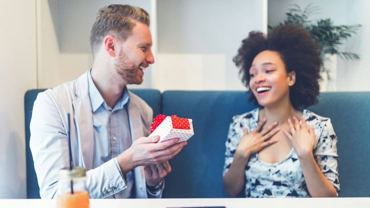 A man smiles as he gives a woman a gift box, and she looks surprised.