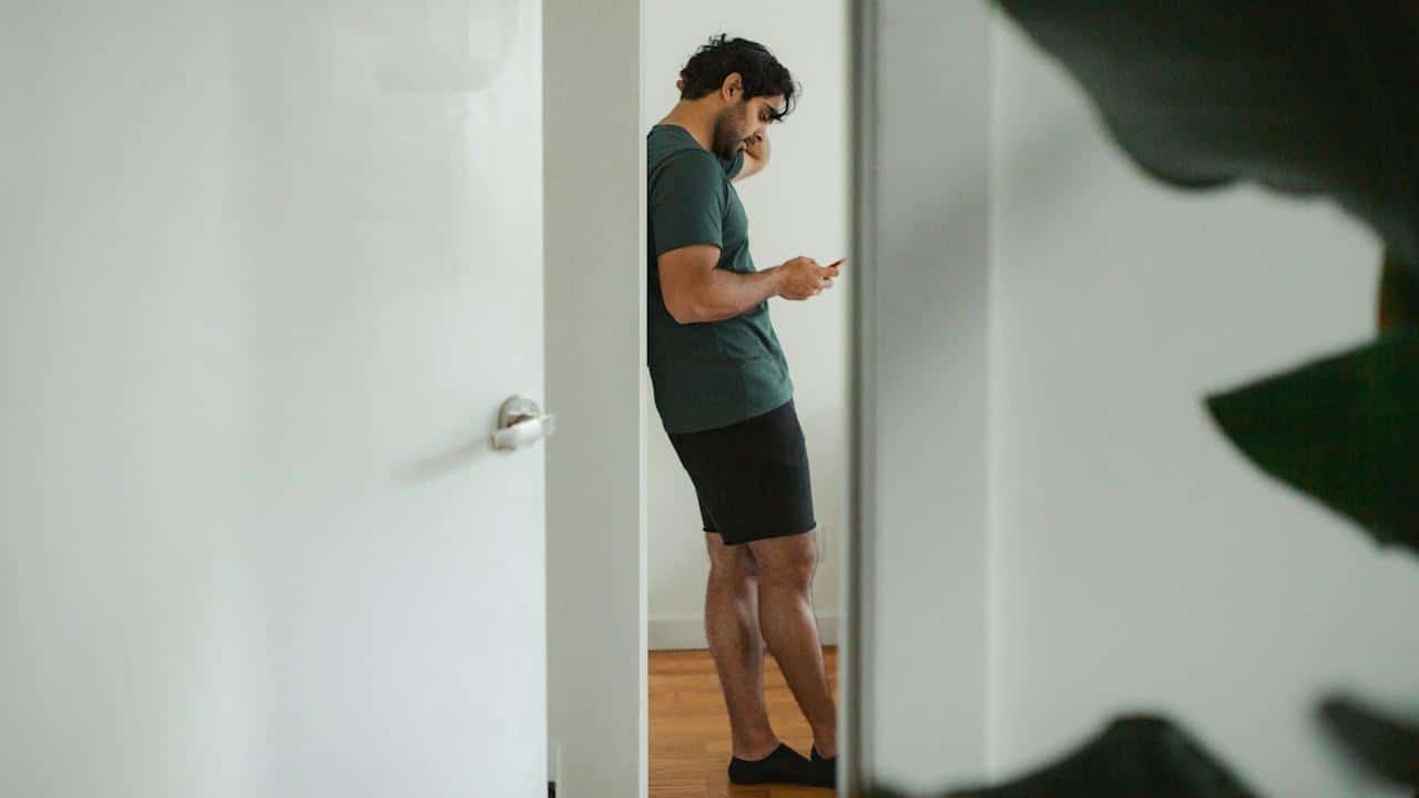 A man in a dark green t-shirt and black shorts stands in a doorway, leaning against the wall, looking down at his smartphone.