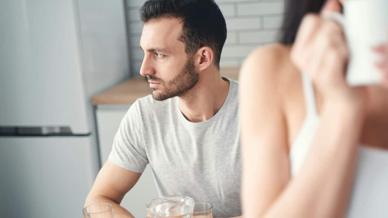Man in a gray shirt sitting at a table in a kitchen, looking away from a person.