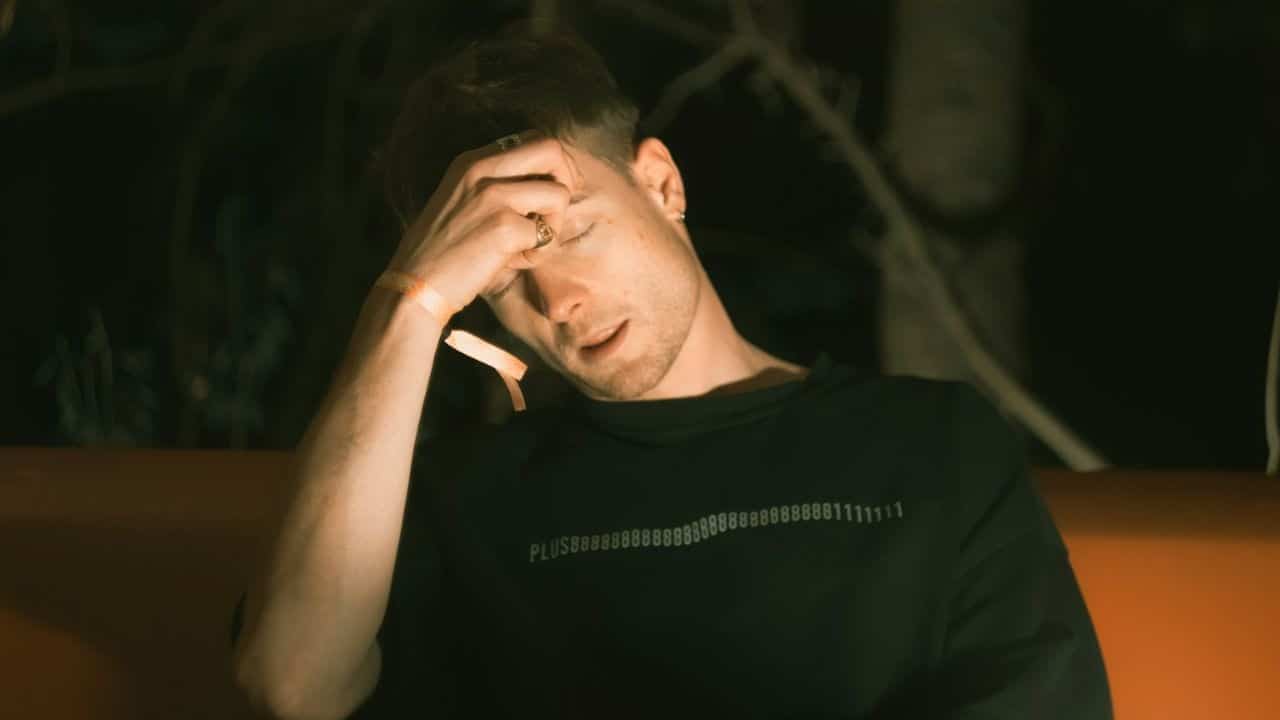 A man sits in the dark with his eyes closed and his hand on his forehead.