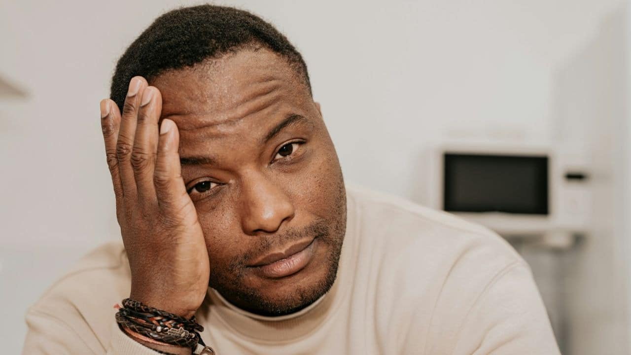 A close-up of a man with his hand on his head, looking directly at the camera.