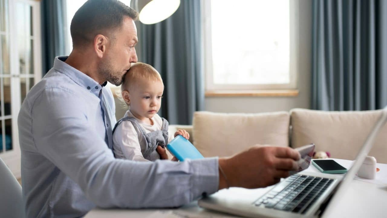 A man holds a baby on his lap while looking at his laptop and holding a phone.