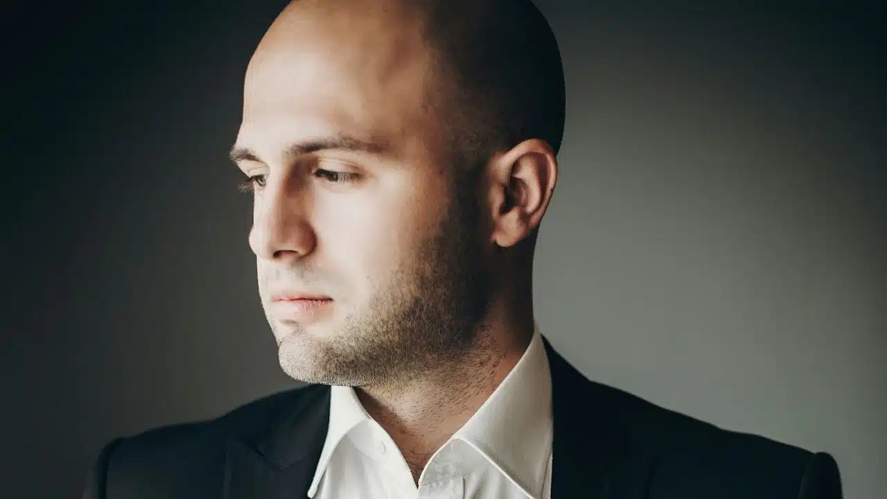 A bald man in a suit and white shirt looks thoughtfully to the side.