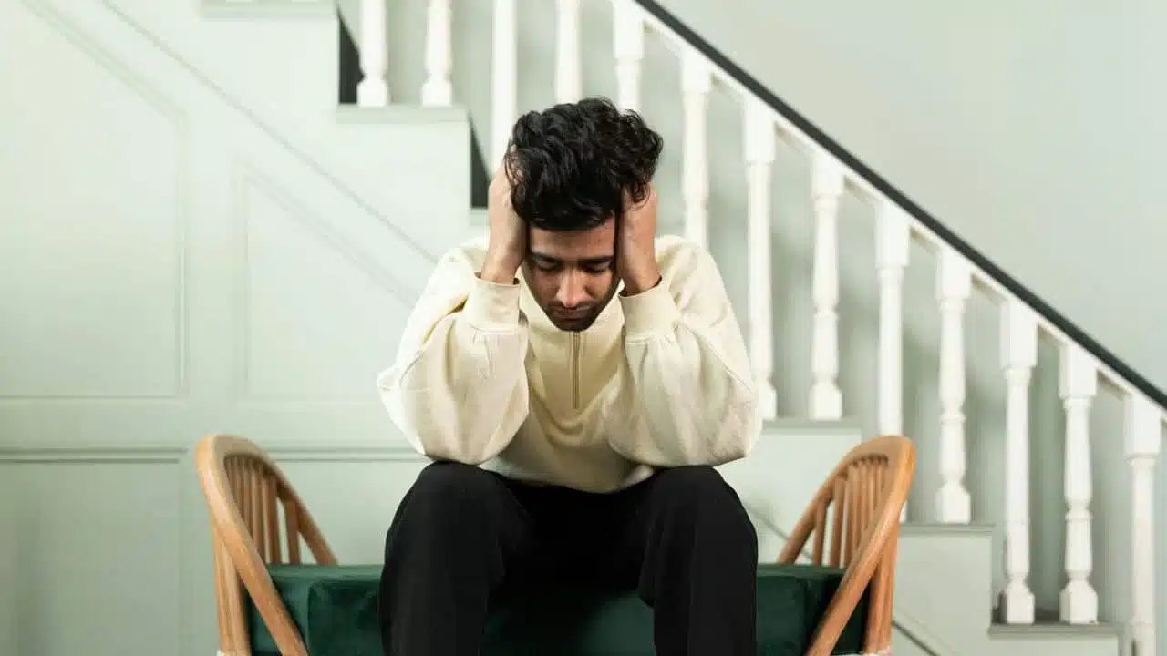 A person sits on a bench, holding their head in their hands, with a staircase in the background.