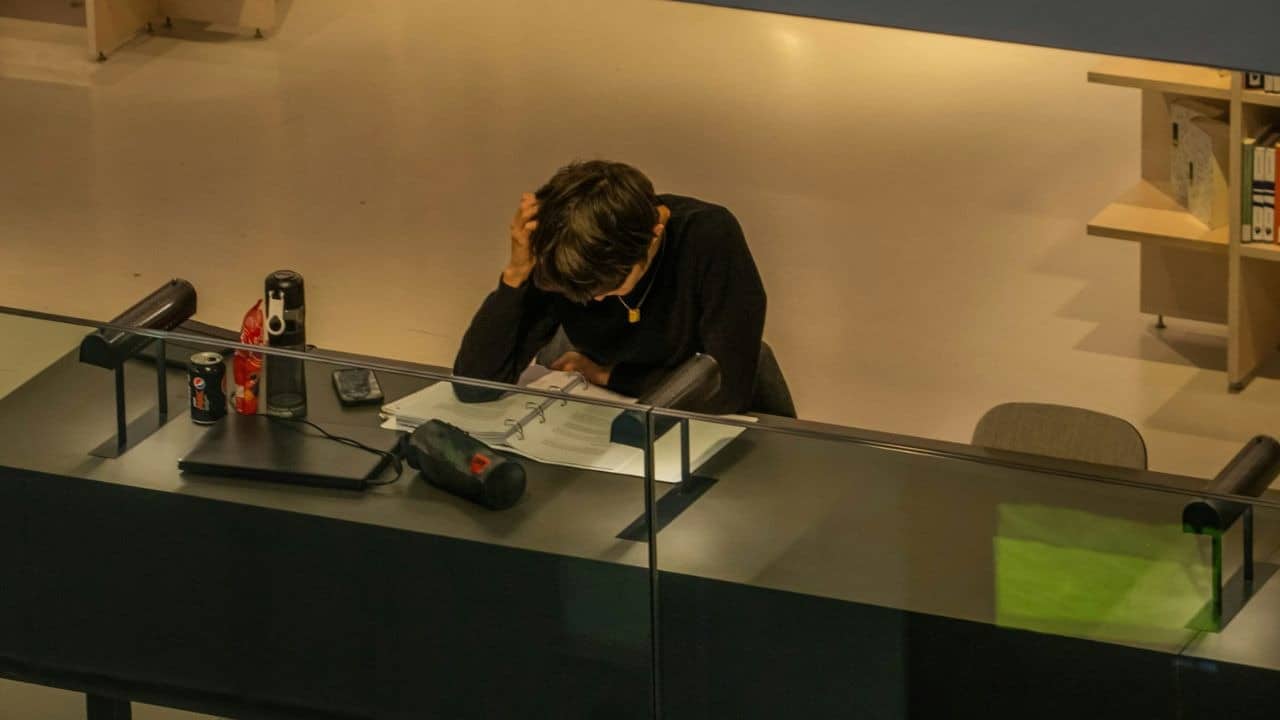 A person with their head in their hand studies at a long desk with a laptop and soda.