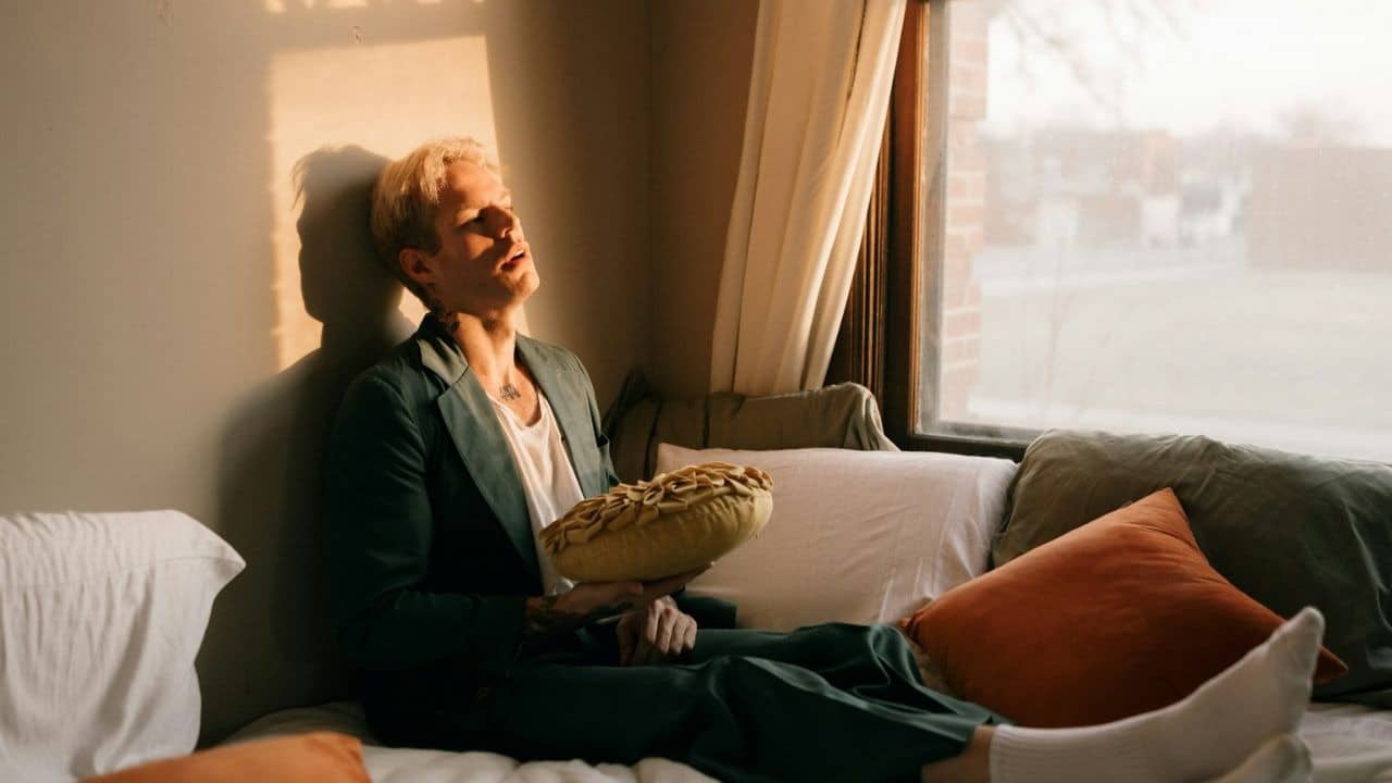 A man in a green suit sits on a cushioned window seat, holding a pie-shaped pillow.