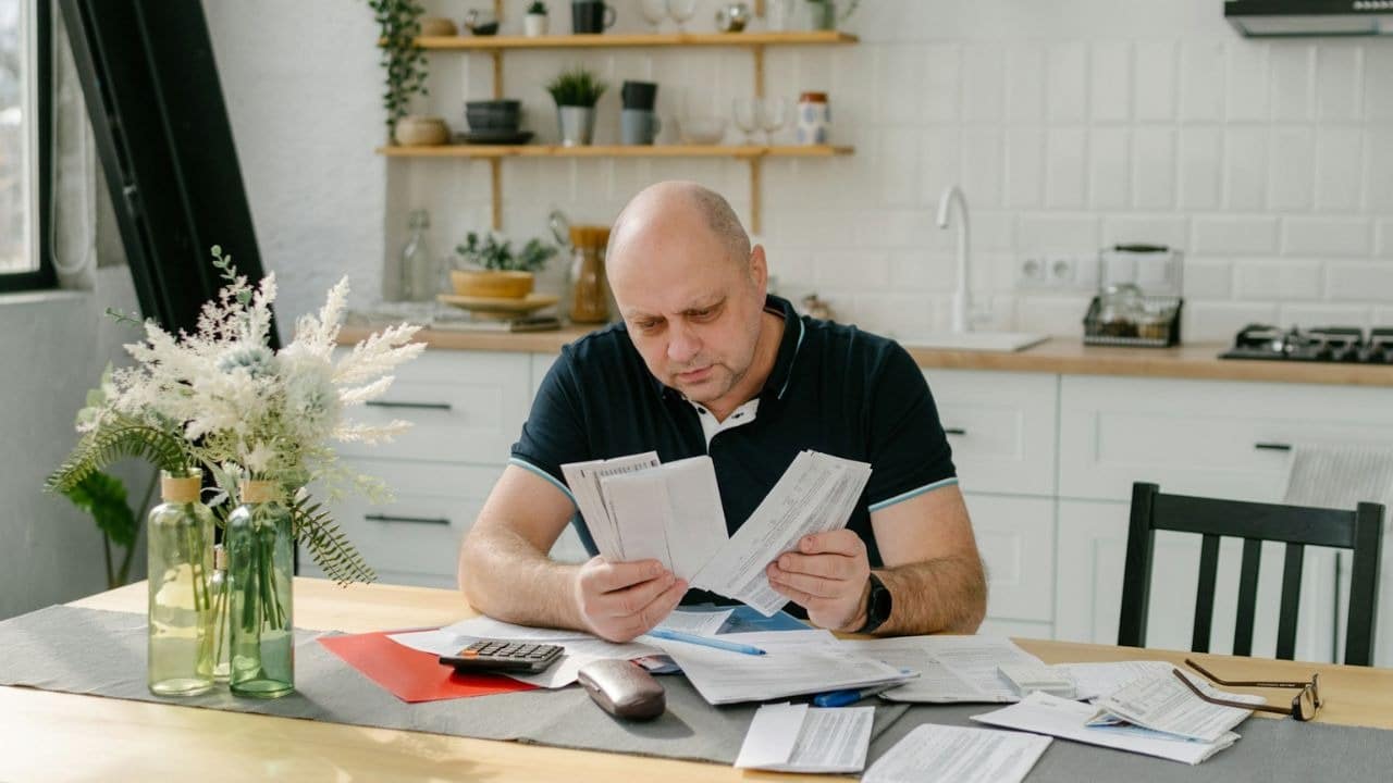 A bald man sits at a kitchen table, looking worried while holding and examining several bills.