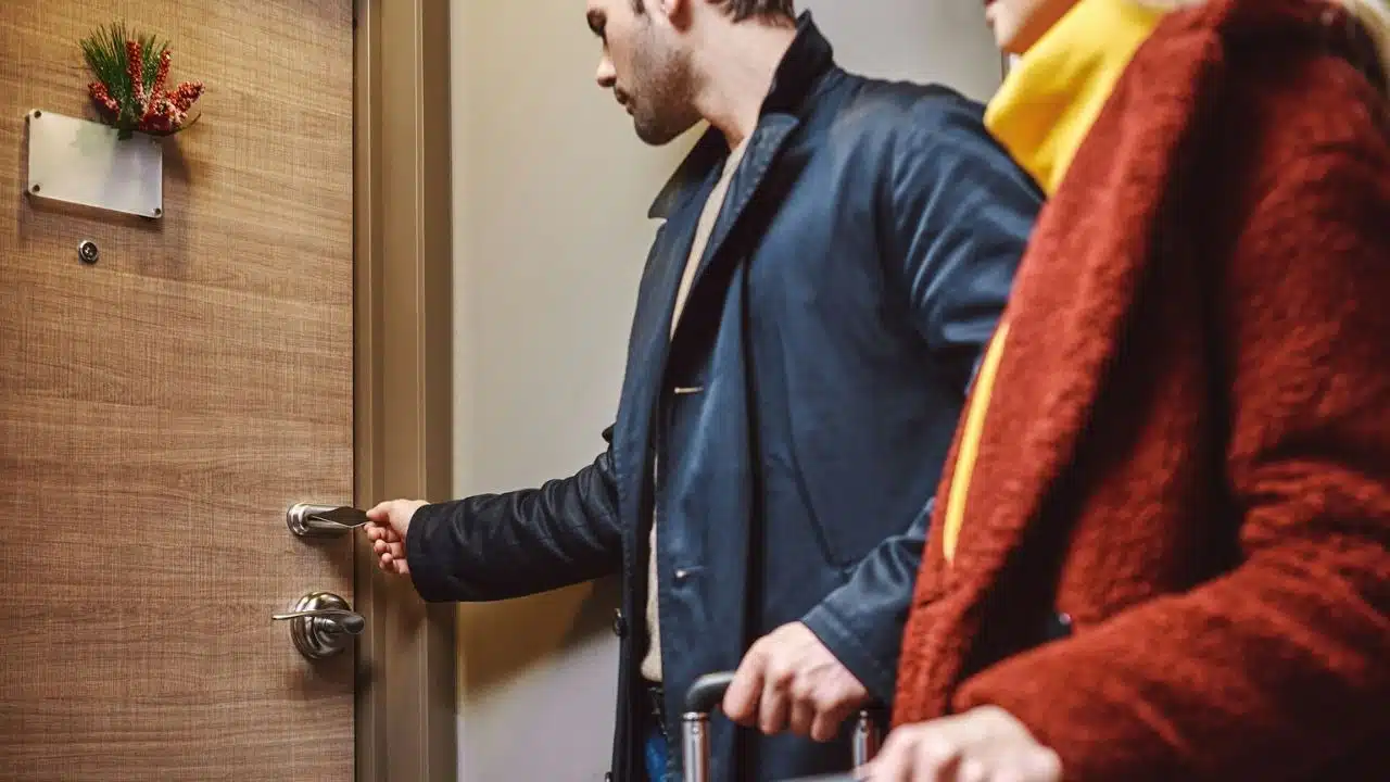 A man in a coat opens a wooden door for a woman with a suitcase.