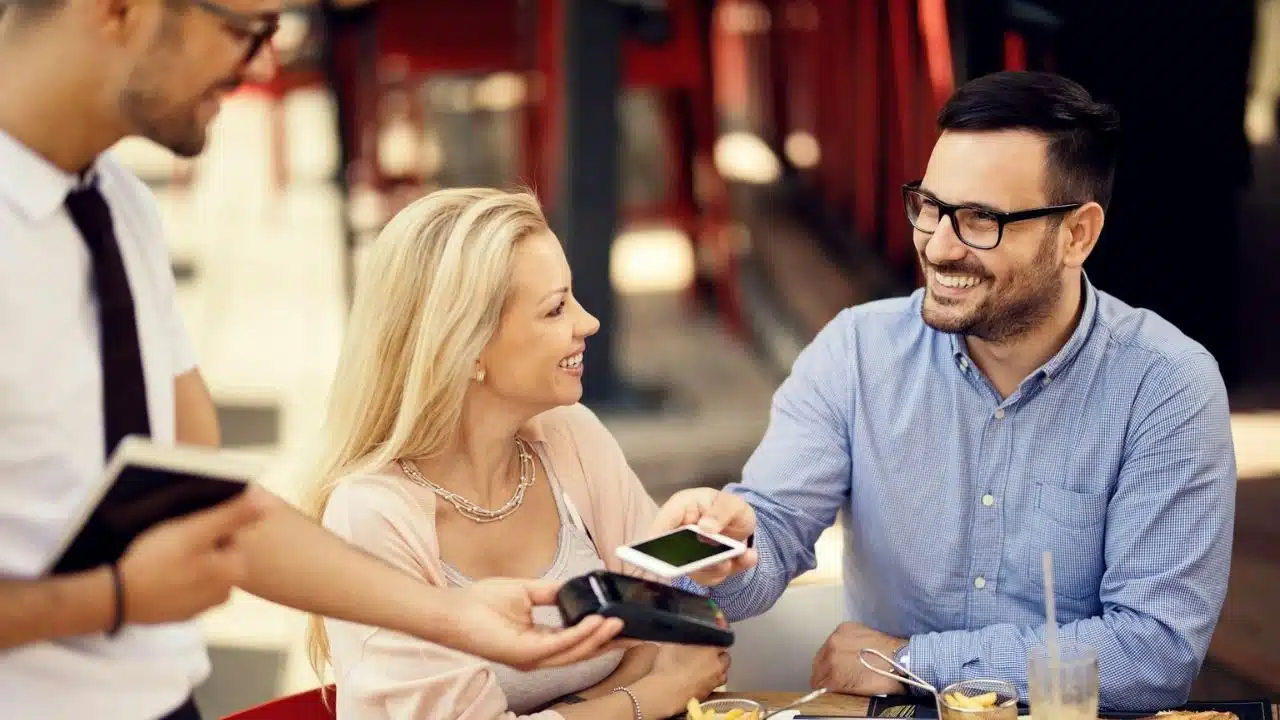 A couple smiles at each other while a man pays with a smartphone at a restaurant.