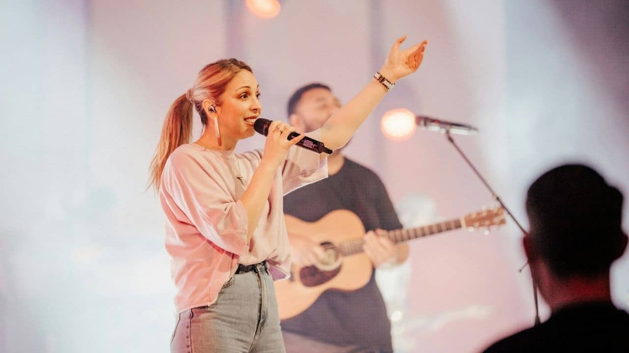 A woman sings into a microphone, with a man playing a guitar in the background.