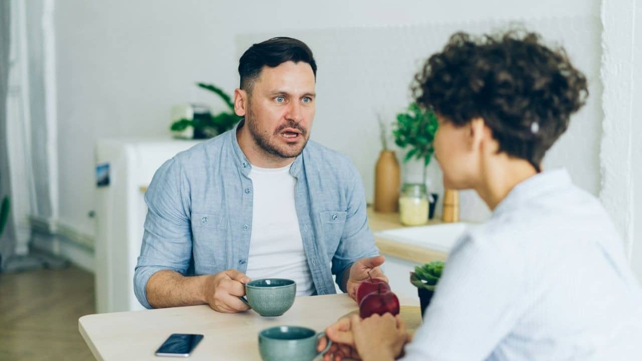 A man talks to a woman at a table, holding a mug.