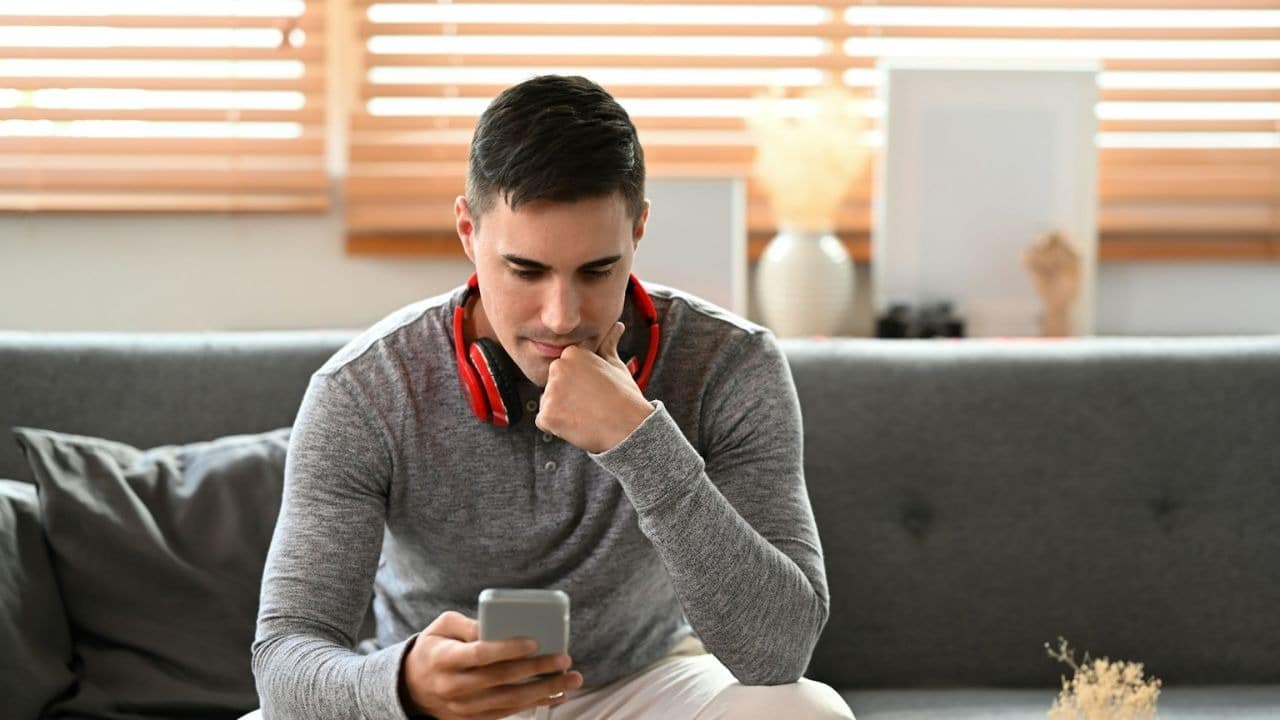 A man with headphones around his neck sits on a couch, looking at his smartphone.