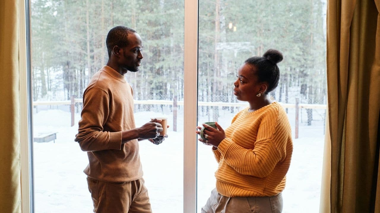 A couple standing by a window, holding cups and talking to one another.