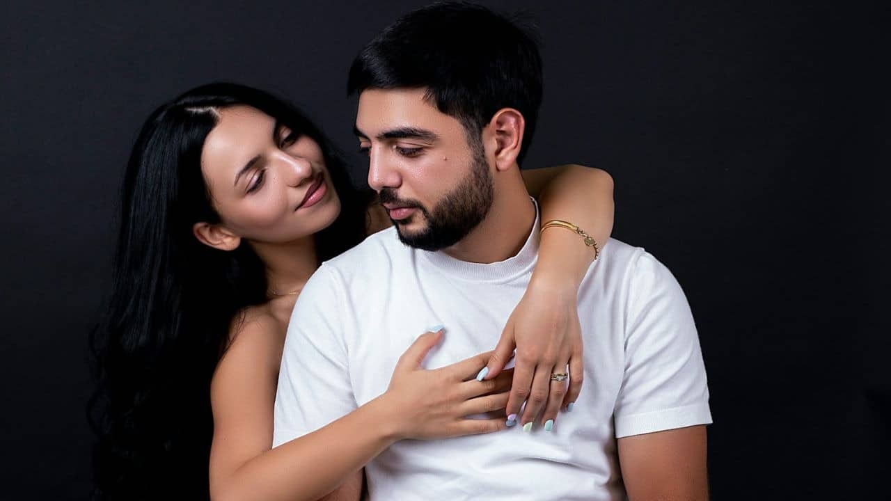 The man is holding the woman's head in his hand, and she is leaning her head against his shoulder.