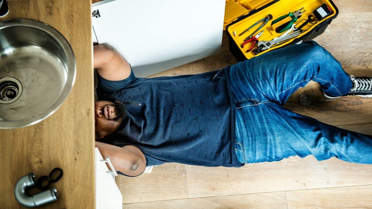 A man is lying on his back under a sink, with an open toolbox nearby.