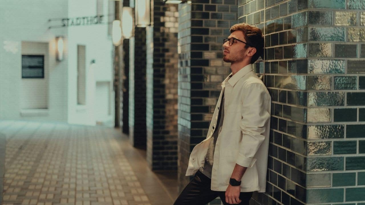 A man with glasses and a white jacket is leaning against a wall with shiny green tiles.
