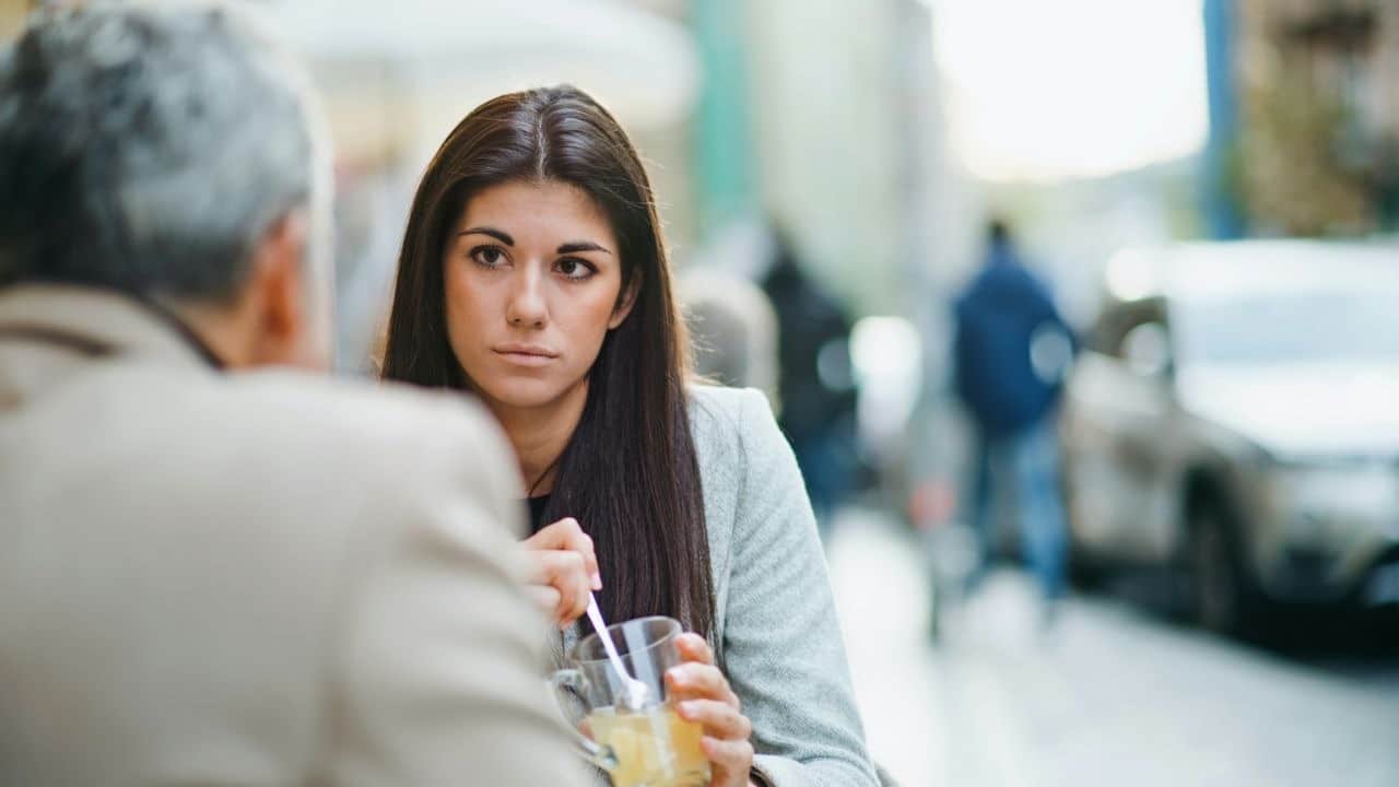 A woman with a serious expression sits at an outdoor cafe, holding a glass.