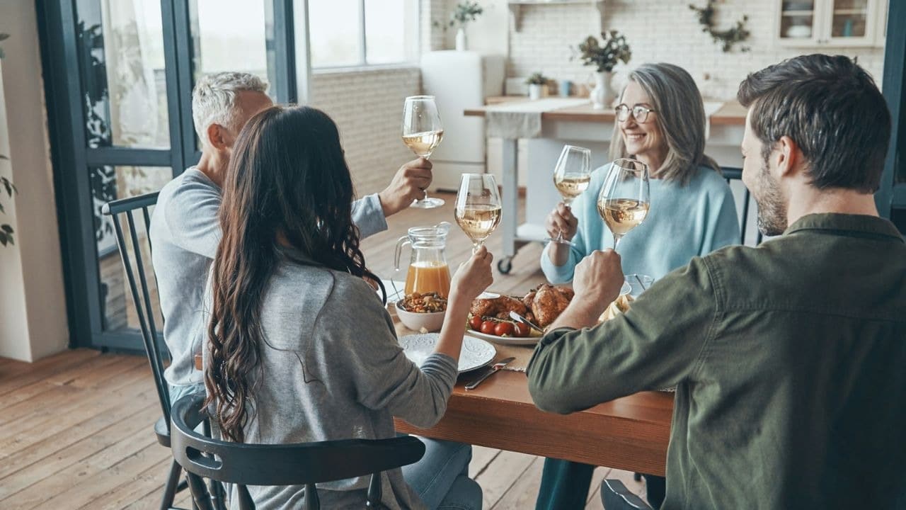 A family of four is seated around a table, holding glasses of white wine and toasting to each other.