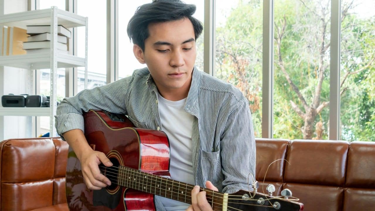 A young man is sitting on a leather couch playing a musical acoustic guitar with a striped shirt.